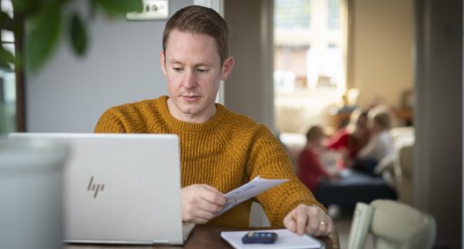 A man - Rob - carries out some admin at a computer, sat at his dining table. A calculate and notepad are at hand.
