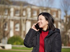 Jacqueline, in remission from DLBCL, smiles whilst holding a phone to her ear, standing outside in front of a house.
