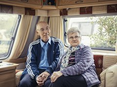 Trevor, who has chronic lymphocytic leukaemia (CLL), sits smiling, holding hands with his wife on the sofa at the back of a caravan.