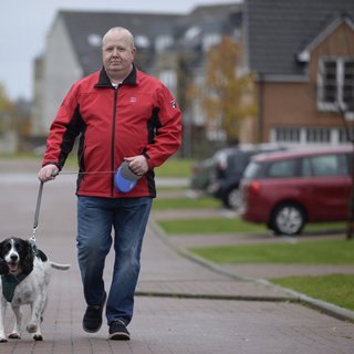 Derek, who is in remission for blood cancer, walks along a street in a modern housing estate with their black and white springer spaniel dog.