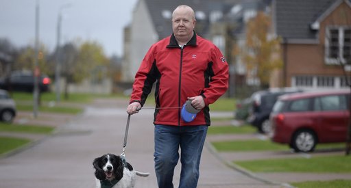 Derek, who is in remission for blood cancer, walks along a street in a modern housing estate with their black and white springer spaniel dog.