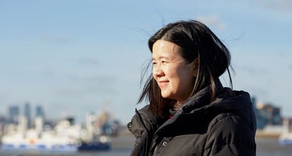 Jacqueline, in remission from DLBCL, stands smiling in the sunshine with a city skyline blurred in the background.