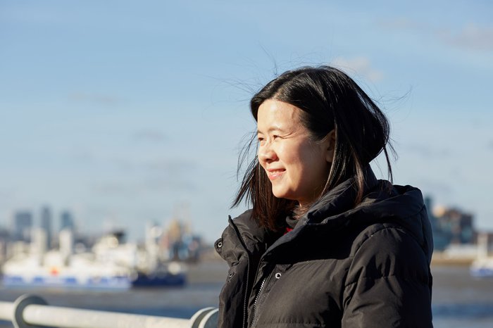 Jacqueline, in remission from DLBCL, stands smiling in the sunshine with a city skyline blurred in the background.