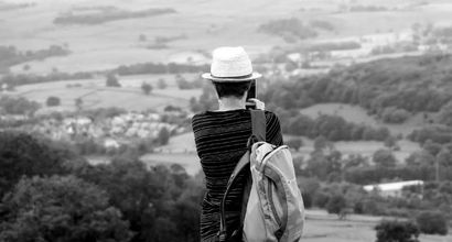 Black and white photo of Becky. Becky is in a field, seen from behind. She's taking a photo on her phone.