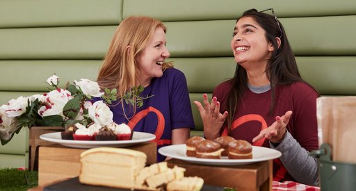 Two women wearing Blood Cancer UK t-shirts laugh together, sitting in front of some cakes.