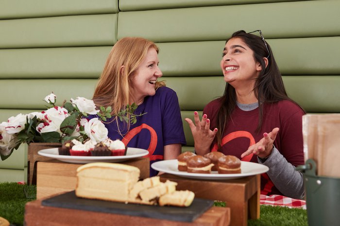 Two women wearing Blood Cancer UK t-shirts laugh together, sitting in front of some cakes.