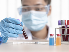 A researcher wearing a facemask inspects a vial of blood on a table with other vials.