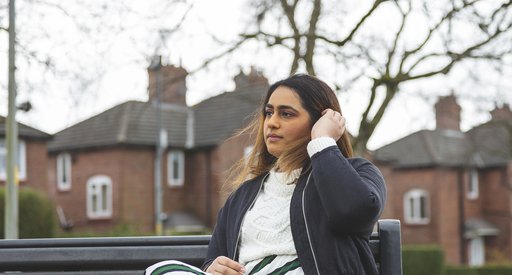 A young woman with blood cancer sits on a bench looking pensive, in front of a row of houses.