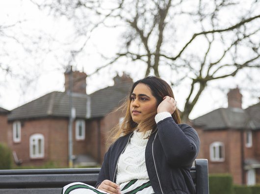 A young woman with blood cancer sits on a bench looking pensive, in front of a row of houses.