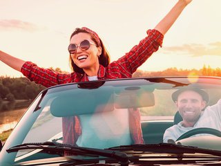 A very happy woman stands up in a convertible car with her hands aloft.