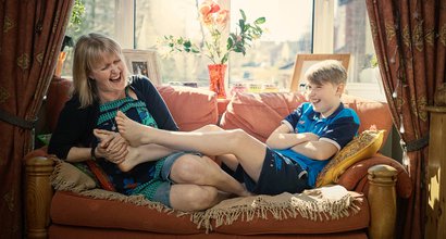 A mother and child laughing together on their sofa at home, the son's feet on his mother's lap.