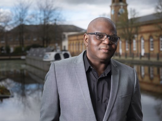 David, wearing a grey suit jacket and black shirt which is open at the neck. Behind David there is a canal boat and building.