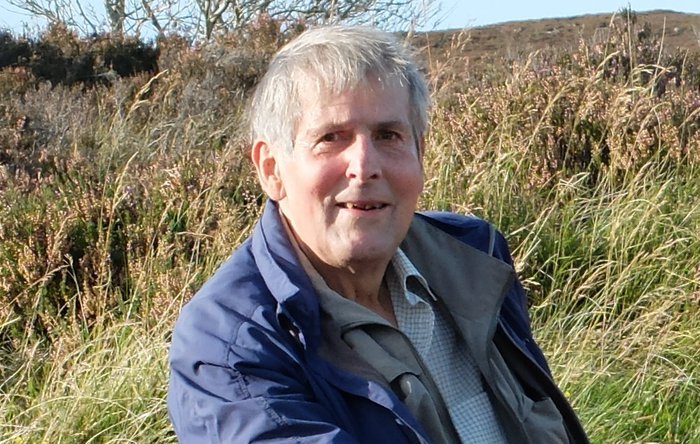 A middle aged man, smiling slightly at the camera, sitting in a green field.