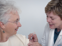 A female doctor in a lab coat injecting a female blood cancer patient at the top of her arm.