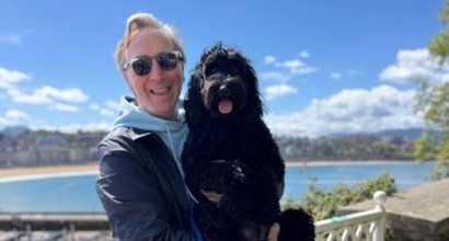 Doug, who had acute myeloid leukaemia, standing on a terrace in the sunshine with a lake or sea in the background. He's smiling, and holds a shaggy black dog in his arms.