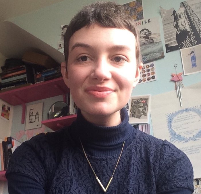 Emma - a young woman - looks straight to camera in a room adorned with posters and books.