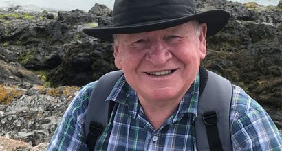 An older man - Gerald - in the countryside. He wears a brimmed hat, check shirt and has a backpack. The image is closely cropped with Gerald looking direct to camera and smiling.