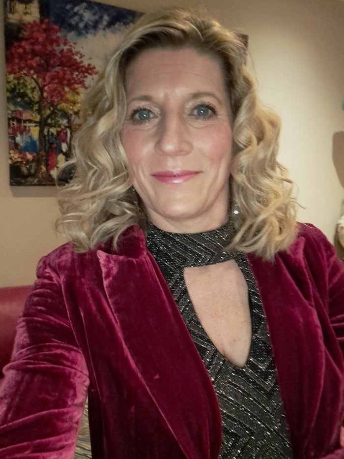 A woman, Helen, wearing a sparkly top and velvet jacket. She is smiling, and there is a painting on the wall behind her.