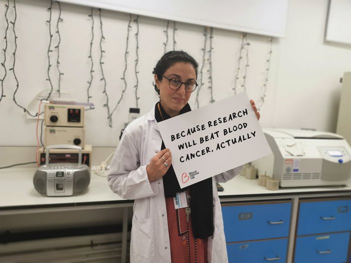 Researcher holding up a sign in a lab which says 'Because research will beat blood cancer, actually' as part of the 2020 Christmas campaign from Blood Cancer UK.
