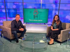 Two presenters sit in a TV studio smiling at the camera, a screen shows the Blood Cancer UK logo