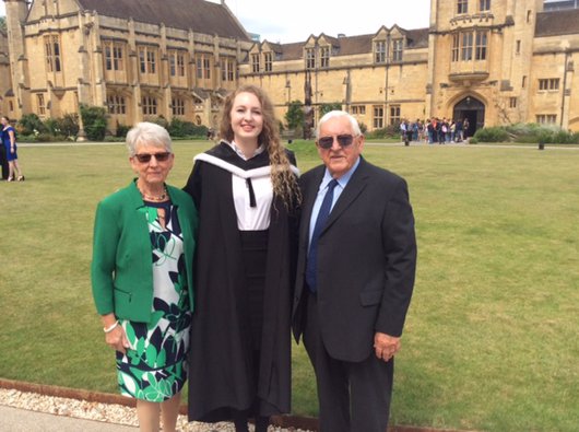 A young graduate wearing a gown, standing with family at her graduation outdoors.