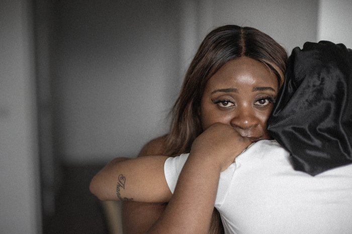 A Black woman hugs another woman in a white t-shirt. She's looking directly into the camera.