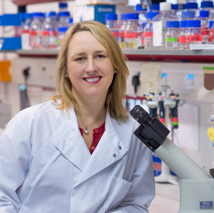 A headshot of Professor Michelle West, wearing a white lab coat and smiling behind a microscope.