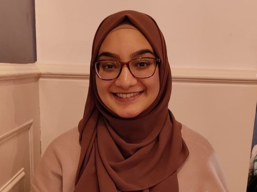 Nabeela, a young woman wearing glasses and a headscarf, smiling at the camera