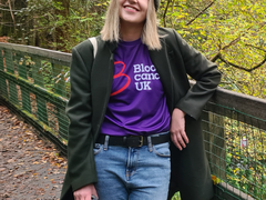 A young woman - Nichola - wearing a Blood Cancer UK t shirt under a winter coat stands smiling on a bridge in the countryside.