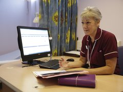 A nurse sits at her desk in a hospital with a computer and notepads.