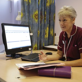 A nurse sits at her desk in a hospital with a computer and notepads.