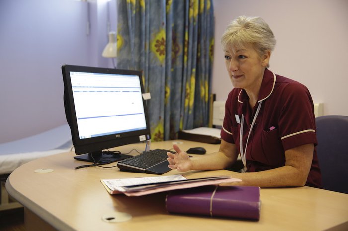 A nurse sits at her desk in a hospital with a computer and notepads.