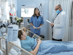 A masked doctor studies a patients chart as he stands over her bed. A nurse also stands nearby.