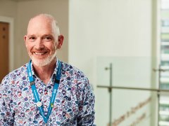 A man stood smiling at the camera wearing a patterned shirt and a blue NHS lanyard.