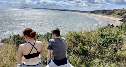 Two young people sitting on a bench, viewed from behind. They're looking out to sea on a sunny day.