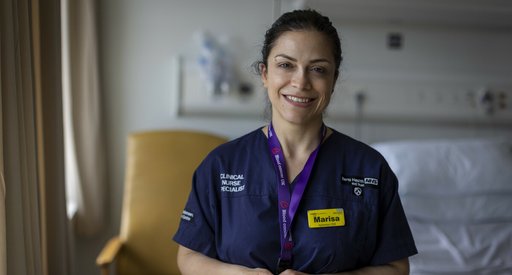 A close up of a healthcare professional wearing blue scrubs, smiling at the camera with hospital equipment out of focus in the background.