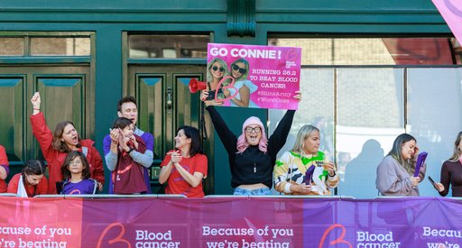 A group of people stand behind partitions at the side of the London Marathon. They are clapping and cheering for the runners taking part in the event. They have clapper boards and a woman with pink hair is holding up a sign that says 'Go Connie!'. It is a sunny day and some of them are wearing Blood Cancer UK t-shirts.