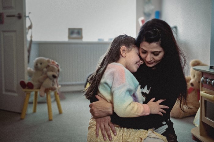 A young girl sits on her mother's lap and gives her a hug. They are sitting in a family home and toys are visible in the background.
