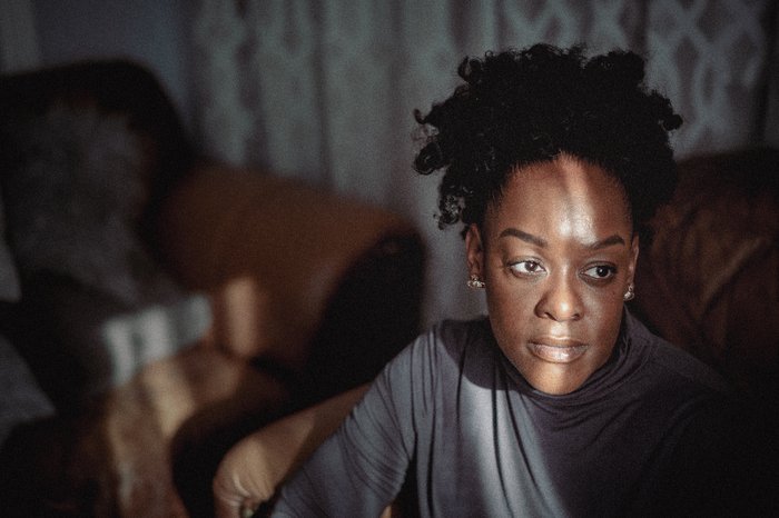 A black woman sits in her family home. She is looking into the distance on her left. There is a ray of light across her face.
