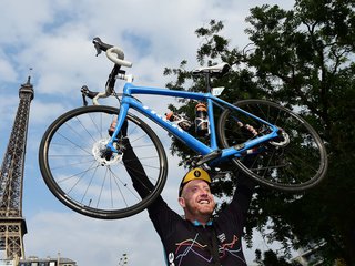 Man in cycling gear smiling whilst holding a blue bike over his head in front of the Eiffel Tower.