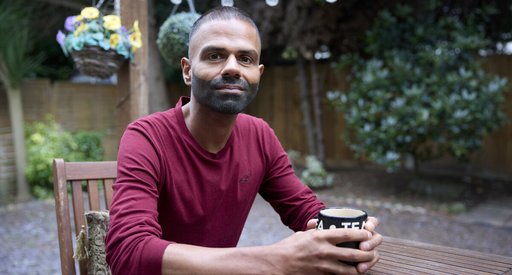 A man is sitting at a garden table, looking at the camera and holding a cup of tea in both hands. He is smiling slightly, and seems thoughtful. There are fairy lights hanging above him and a basket of flowers in the background.