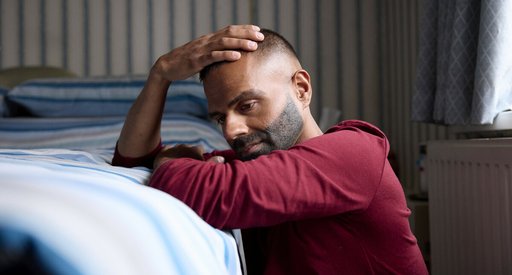 A man on the floor, leaning his elbows on the top of his bed, with one hand placed on the top of his head, looking despondent.