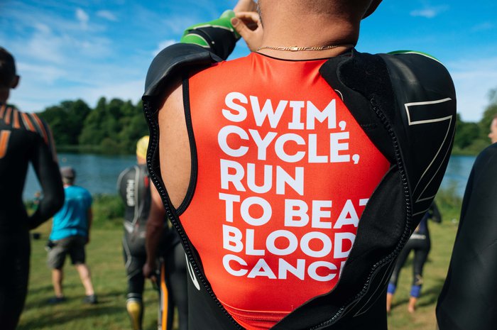 Back of a man by a lake putting on a wetsuit with a Blood Cancer UK swim top underneath.