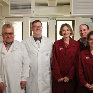 Four researchers in a lab in Kings College London. They are wearing burgundy Blood Cancer UK lab coats and smiling.