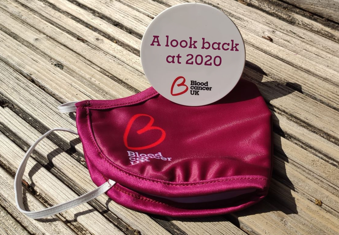 A close up of a Blood Cancer UK face mask and a Blood Cancer UK badge on a wooden table.