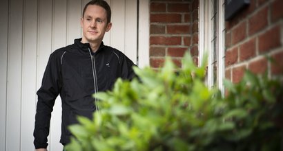 A man stands outside a brick house with a garage door in the background and a plant in the foreground.