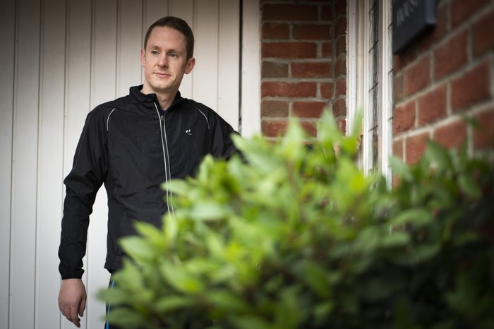 A man stands outside a brick house with a garage door in the background and a plant in the foreground.