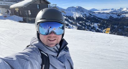 Russell is standing on a snow-covered mountain, smiling at the camera. He is holding a snowboard and wearing ski goggles.