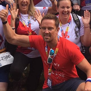 Simon Thomas, President of Blood Cancer UK, kneeling with his right arm raised in front of a crowd of people posing and cheering at an event.