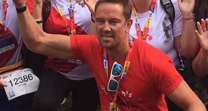 Simon Thomas, President of Blood Cancer UK, kneeling with his right arm raised in front of a crowd of people posing and cheering at an event.